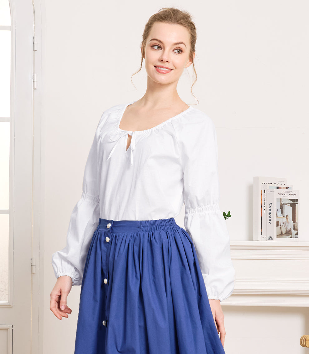 Indoor portrait of a woman wearing an ivory vintage aesthetic blouse and a blue buttoned skirt standing next to a white fireplace.